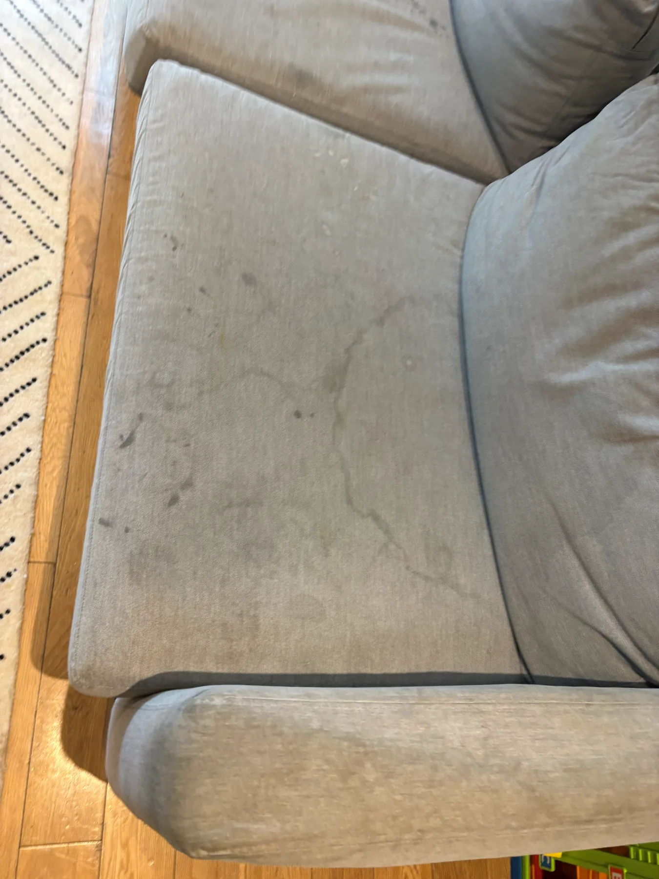 A taupe fabric sofa seat cushion before cleaning, marked with dark patches and the dried rings of old spills