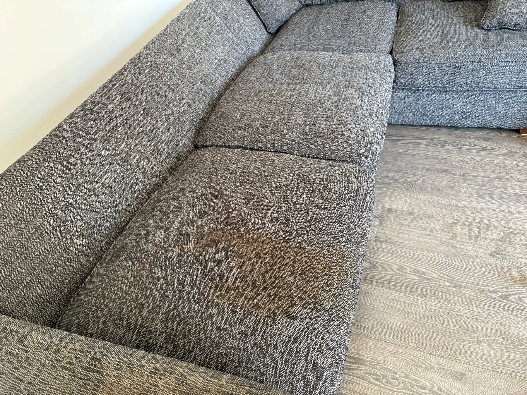 A grey corner sofa before cleaning, with a large darkened patch worn into the middle of the chaise seat