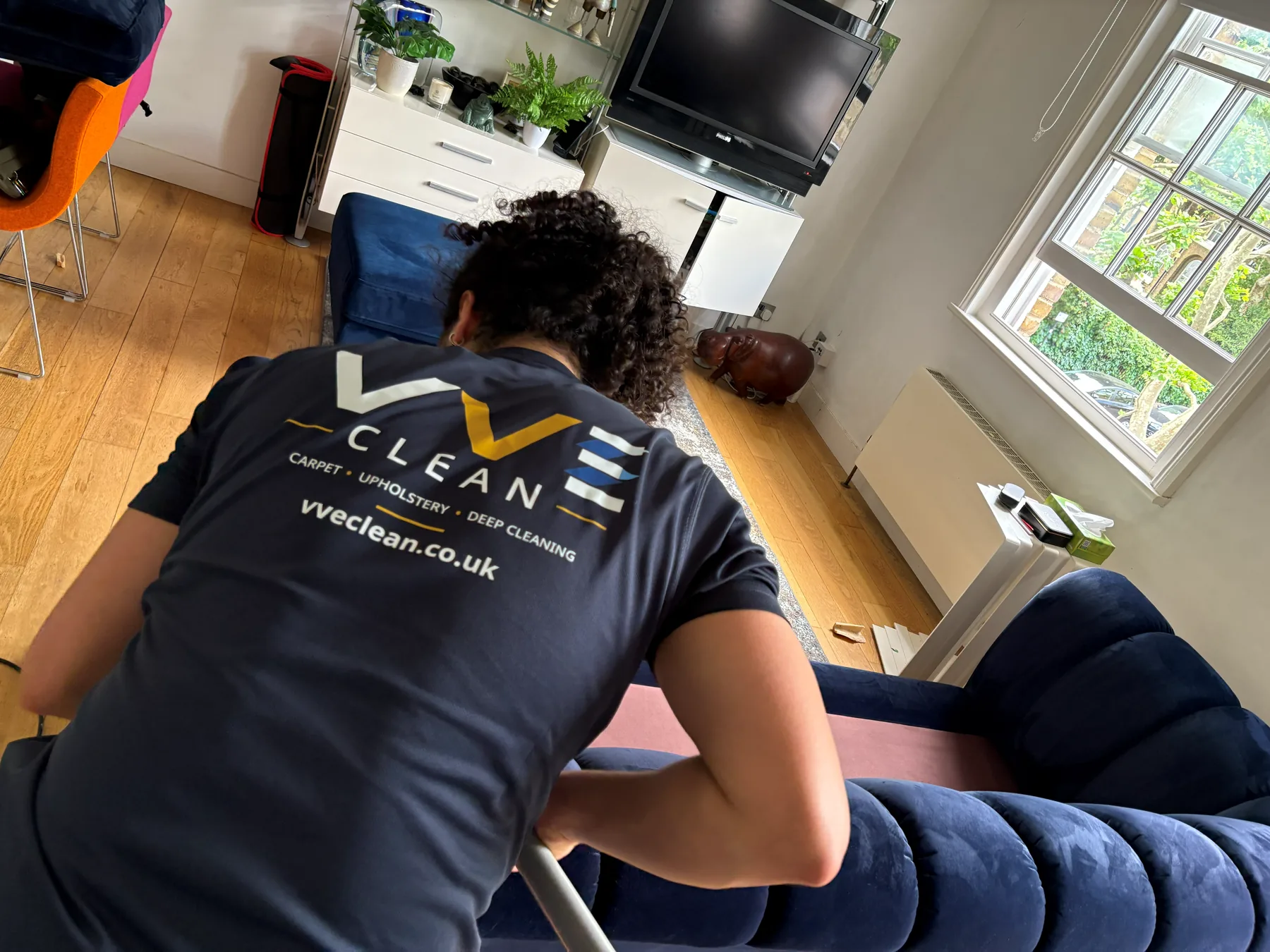 A VVE Clean technician in branded uniform leaning over a navy velvet sofa mid-clean in a bright living room