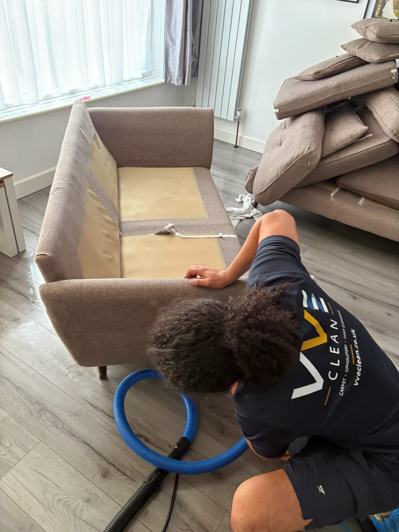 A VVE Clean technician kneeling with the blue extraction hose beside a taupe sofa stripped of its cushions