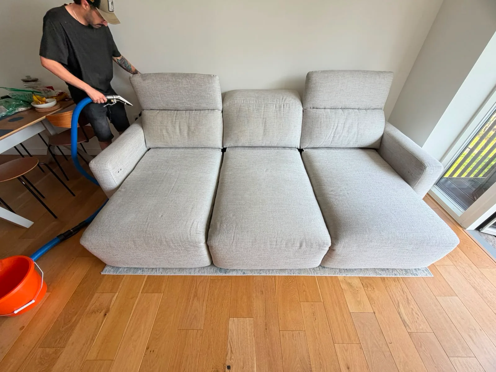 A technician working a hand extraction tool across the back of a pale grey three-seater sofa beside a patio door